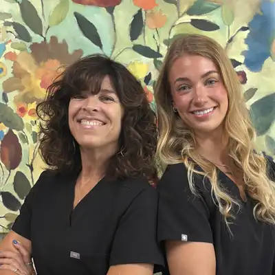 Two chiropractic clinic team members in black scrubs posing in front of a colorful floral wall art display inside a healthcare office
